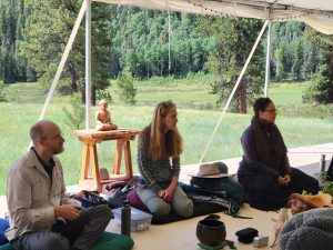 3 Guiding Teachers sit in a large tent facing a group of retreatants. A field of grass, trees and mountains are in the background behind the teachers..